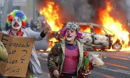 Demonstrators dressed as clowns pass by a burning police car during the Frankfurt protests. / Michael Probst/AP
