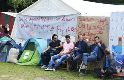 A group of asylum seekers resting at mid-day. Behind them the slogan says: “Without papers and refugees are the same: a problem of papers”. / J. Philippe