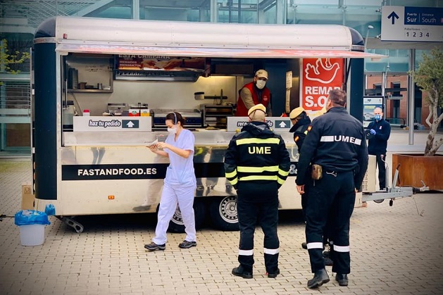 A nurse picks up her lunch from the REMAR food stand located at the entrance of the IFEMA, as Military Emergencies Unit (known as UME in Spanish) personnel wait their turn. / Remar via Actualiada Evangélica,