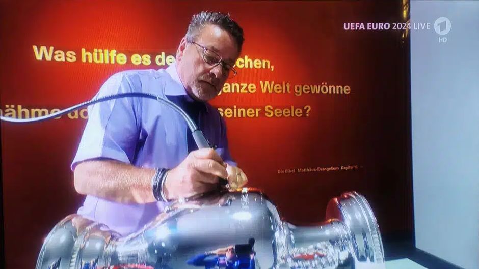 The European Championship trophy is being engraved and the Bible verse can be seen in the background. / Photo via <a target="_blank" href="https://www.pro-medienmagazin.de/"> Pro Medien Magazin </a>.,